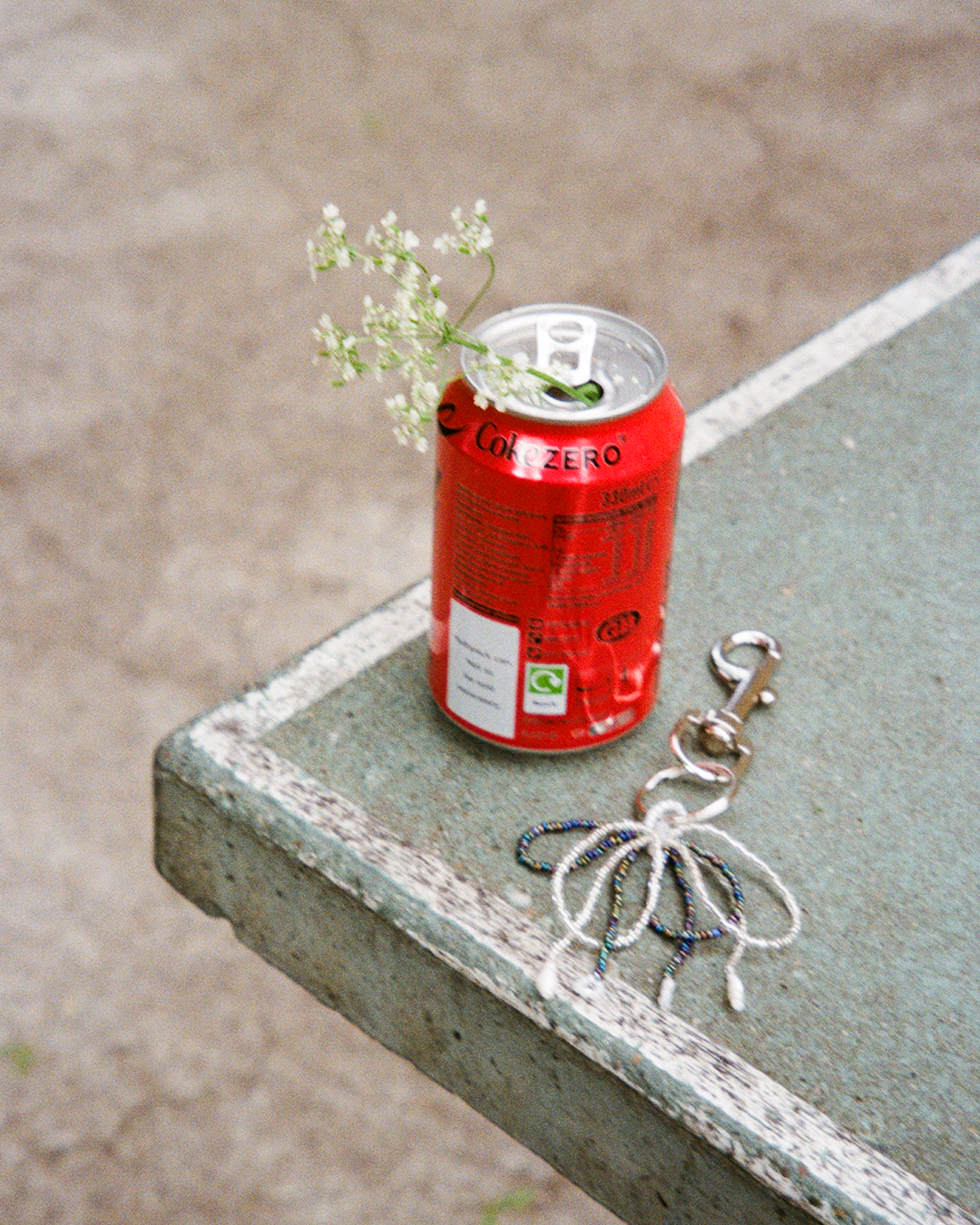 Dark beaded bow keychain on silver clip with dangling pearl and flower charms next to a can of soda.