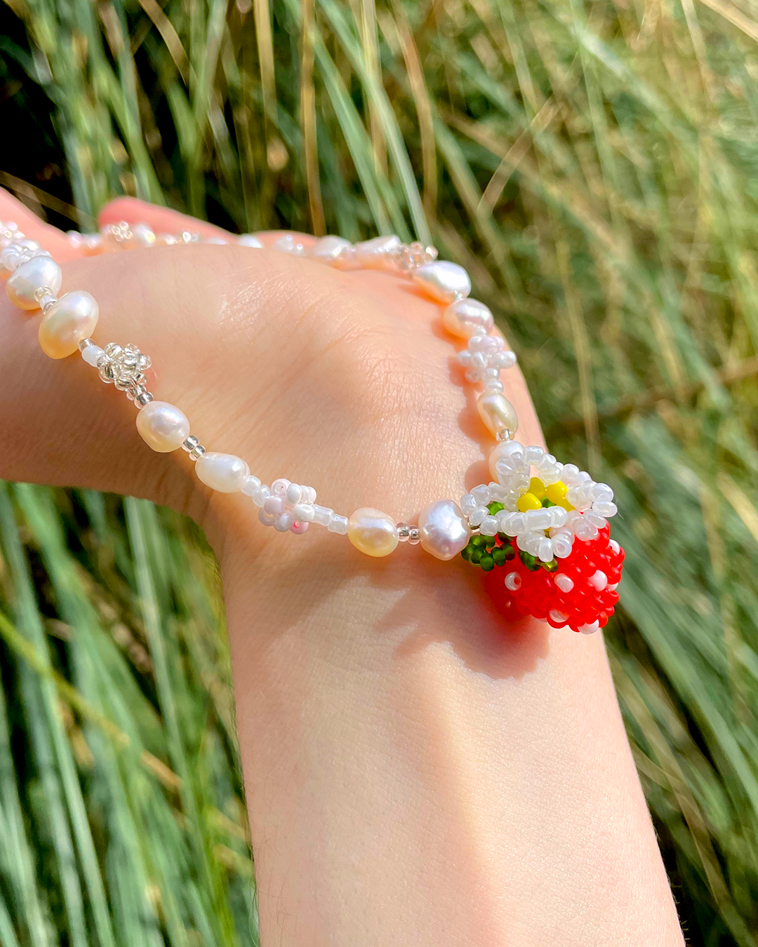 Hand showing a beaded necklace with strawberry charm against a grassy background