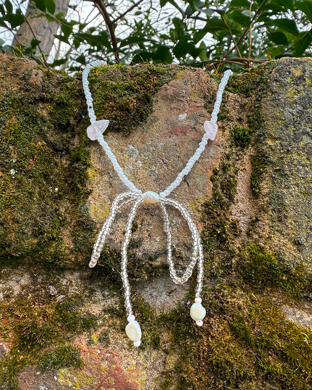Necklace featuring a clear beaded bow centrepiece with hanging rose quartz and pearl charms hanging on a mossy wall.