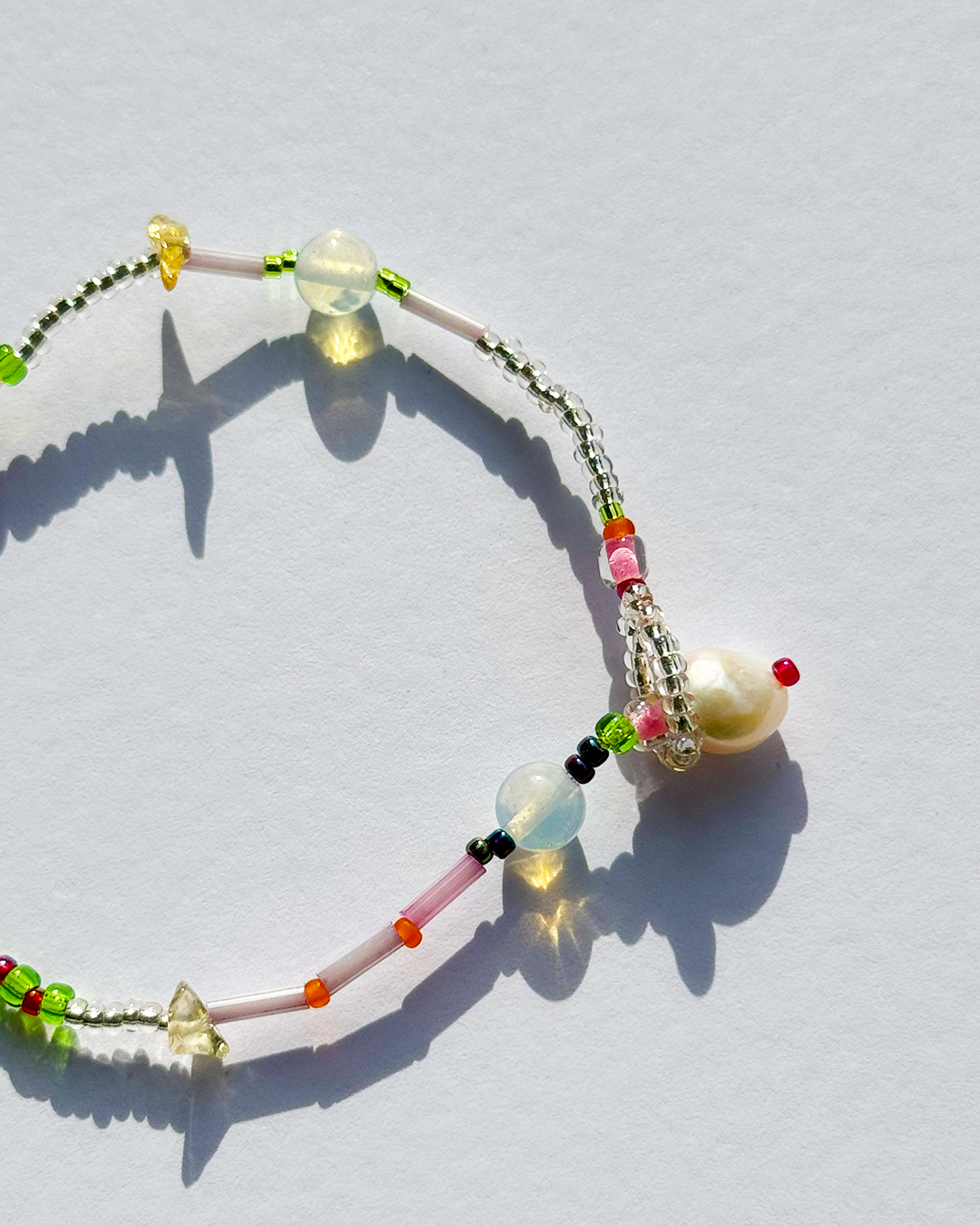A delicate beaded bracelet with a large white pearl bead, featuring translucent light blue, pink, and green beads, silver-toned seed beads, and small colorful accent beads, arranged on a white surface with sharp shadows.