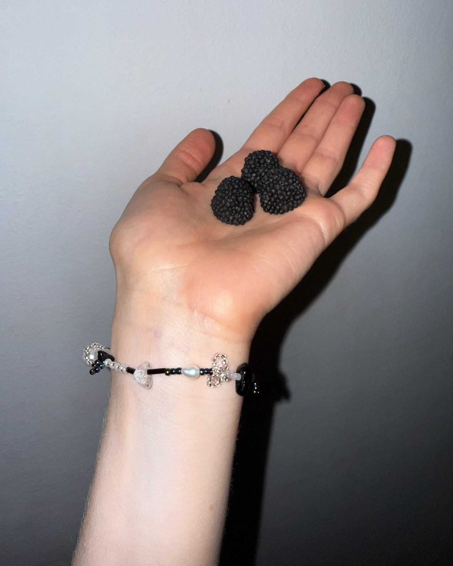 A person's hand holding three blackberries, wearing a beaded bracelet on the wrist. The bracelet features black, white, and clear beads with silver-toned charms.