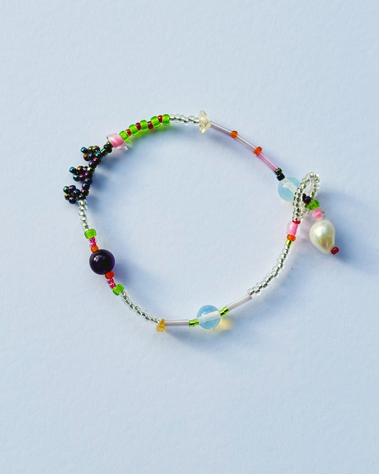 A delicate beaded bracelet with a silver-toned chain, featuring green, pink, red, and blue beads, a dark purple bead, a small white flower charm, and a dangling white pearl, displayed on a light blue background.
