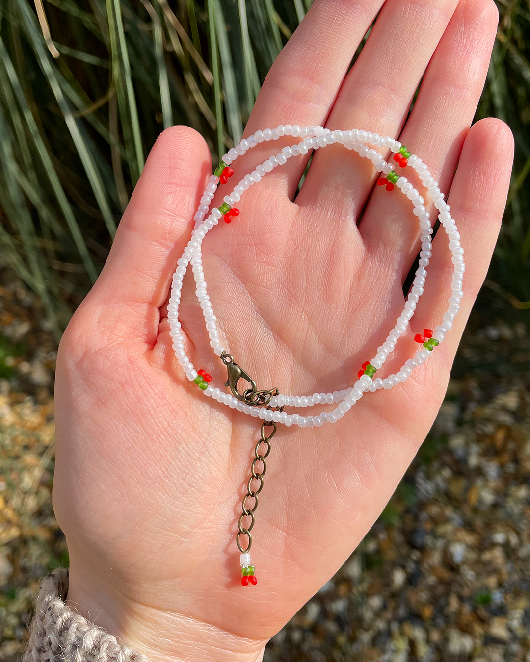 Hand holding a delicate beaded necklace with red and green accents against a natural background.