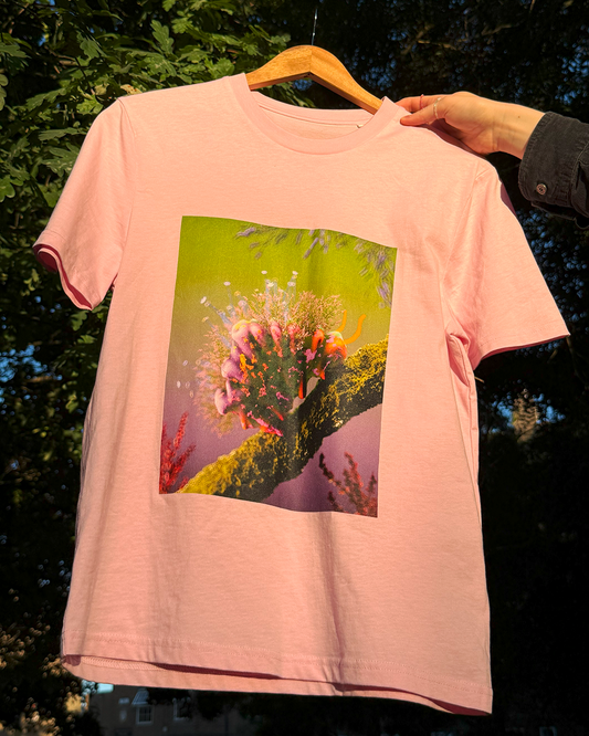 Pink t-shirt with a square print featuring a fuzzy, colorful fantasy caterpillar 3D artwork, against natural background.