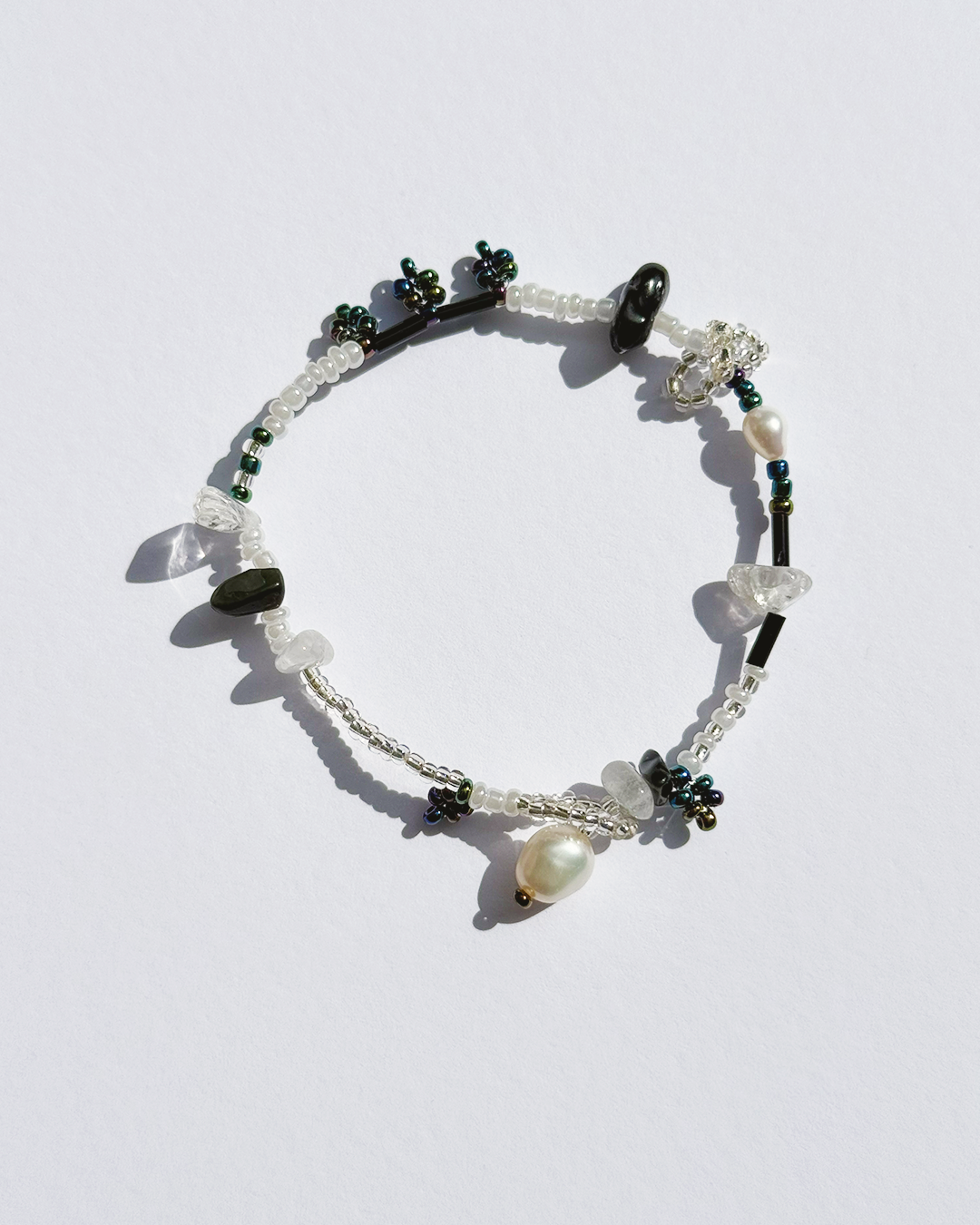 A delicate beaded bracelet with white translucent crystal beads, dark green faceted beads, a white pearl, and a silver-toned toggle clasp, displayed on a light white background.