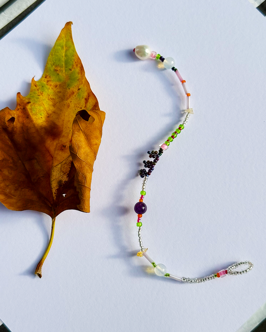 A beaded  bracelet with silver chain and colorful beads, styled on a white surface next to a brown autumn leaf.