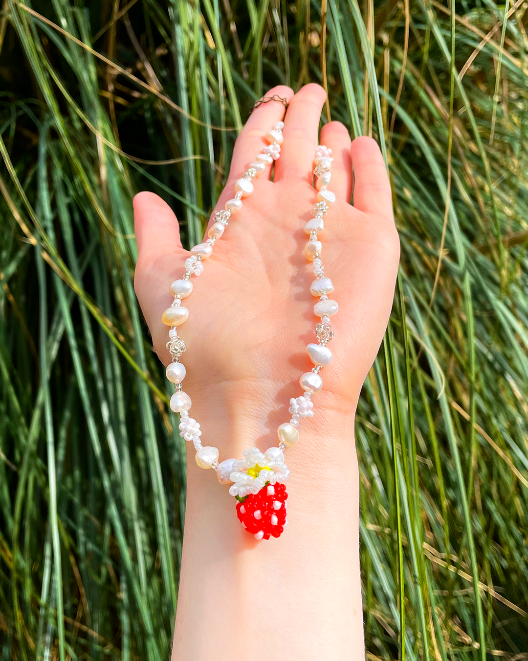 Hand showing a necklace with a red strawberry charm against a grassy background