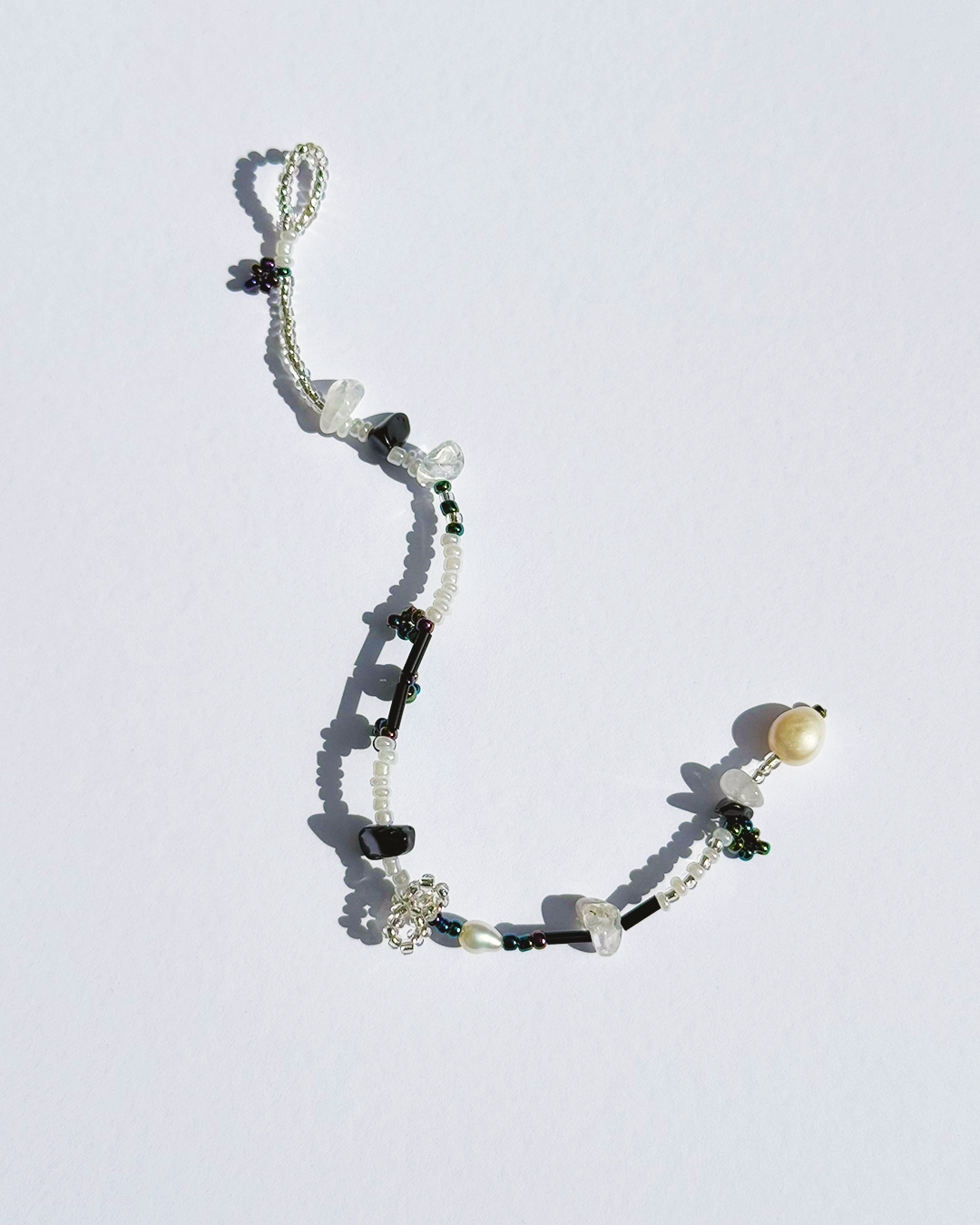 A delicate beaded bracelet with black and white beads, clear crystal beads, a white pearl, and silver-toned floral charms, displayed on a light white background.
