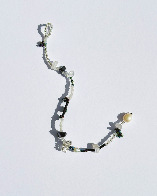 A delicate beaded bracelet with black and white beads, clear crystal beads, a white pearl, and silver-toned floral charms, displayed on a light white background.