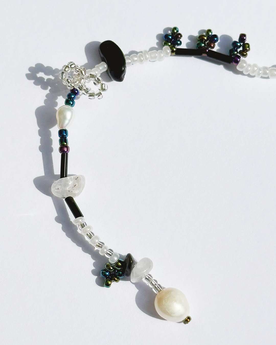 A beaded bracelet with white pearls, black and clear crystal beads, and small iridescent green and blue beads, arranged on a white background.