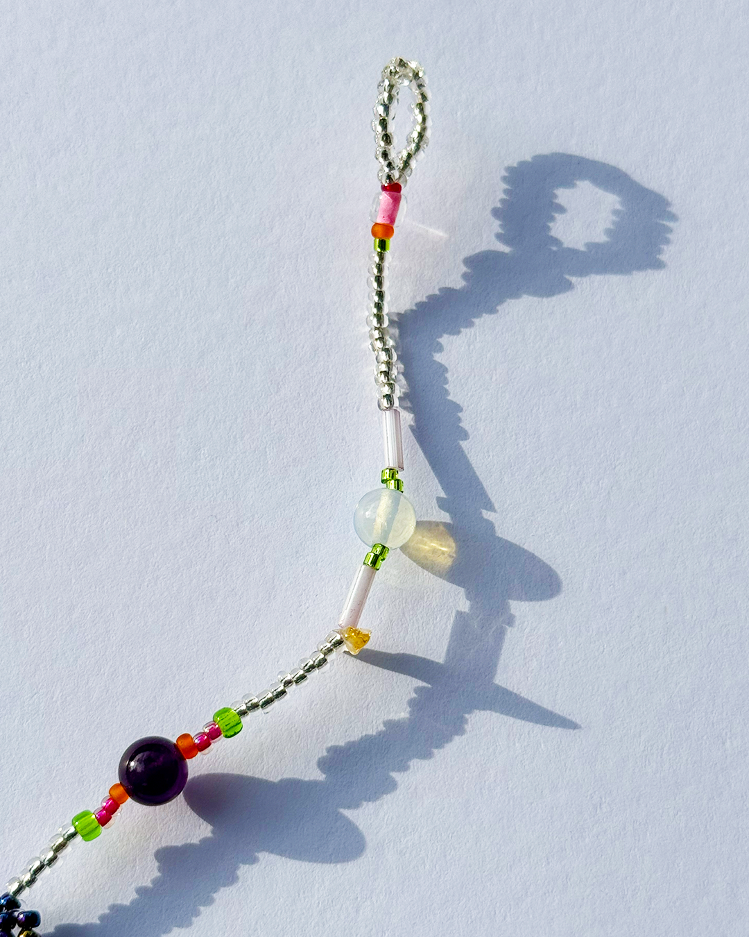 A beaded bracelet with a silver-toned beaded loop, featuring white, green, orange, red, and purple beads, displayed on a light blue surface with a sharp shadow.
