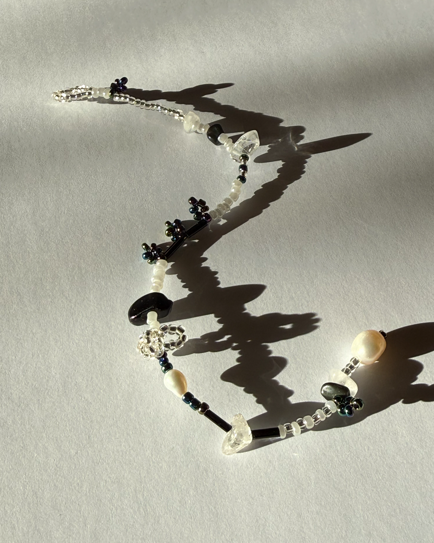 A beaded bracelet with white pearls, clear faceted beads, and dark blue floral accents, arranged on a light gray surface with strong shadows.