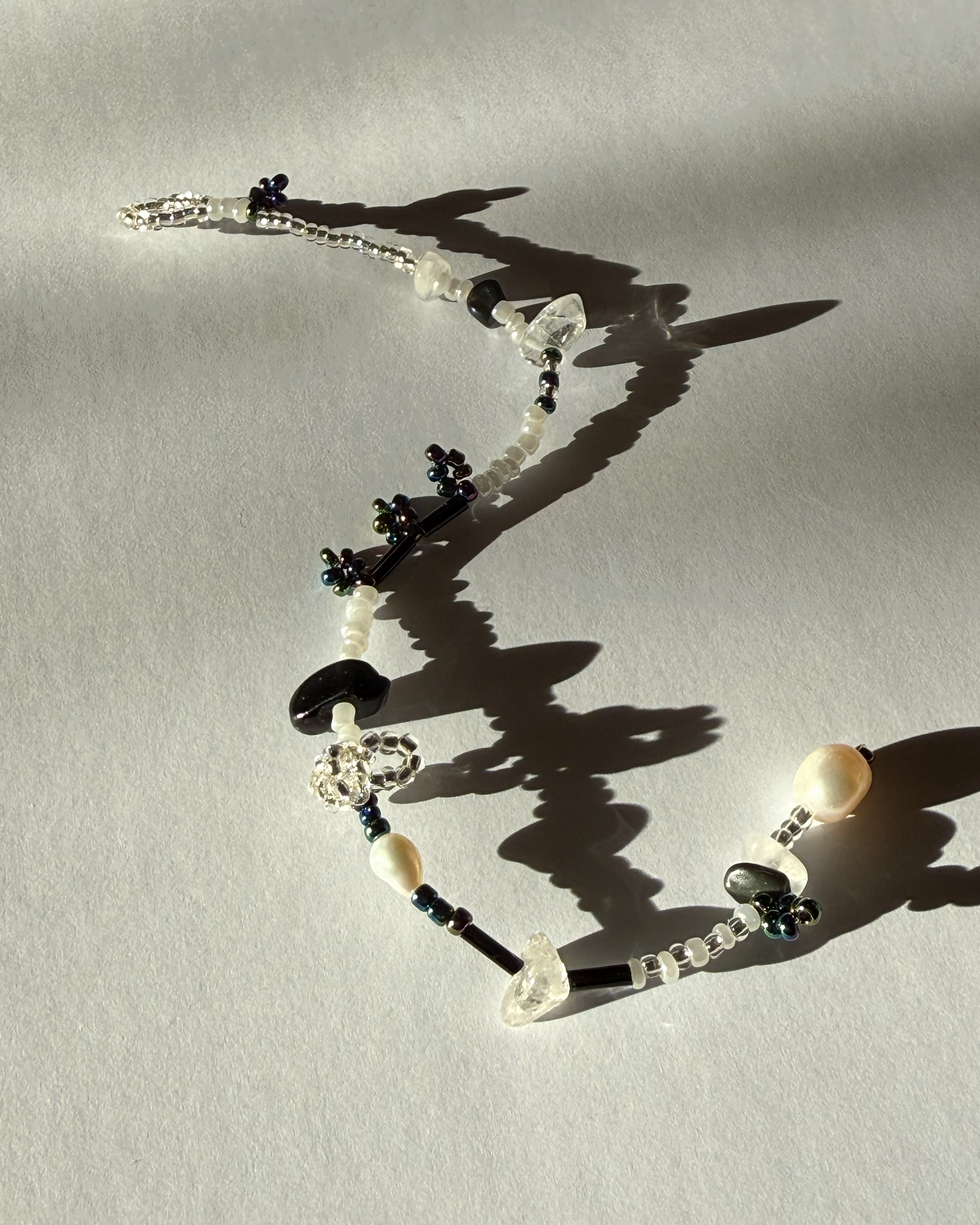 A beaded bracelet with white pearls, clear faceted beads, and dark blue floral accents, arranged on a light gray surface with strong shadows.