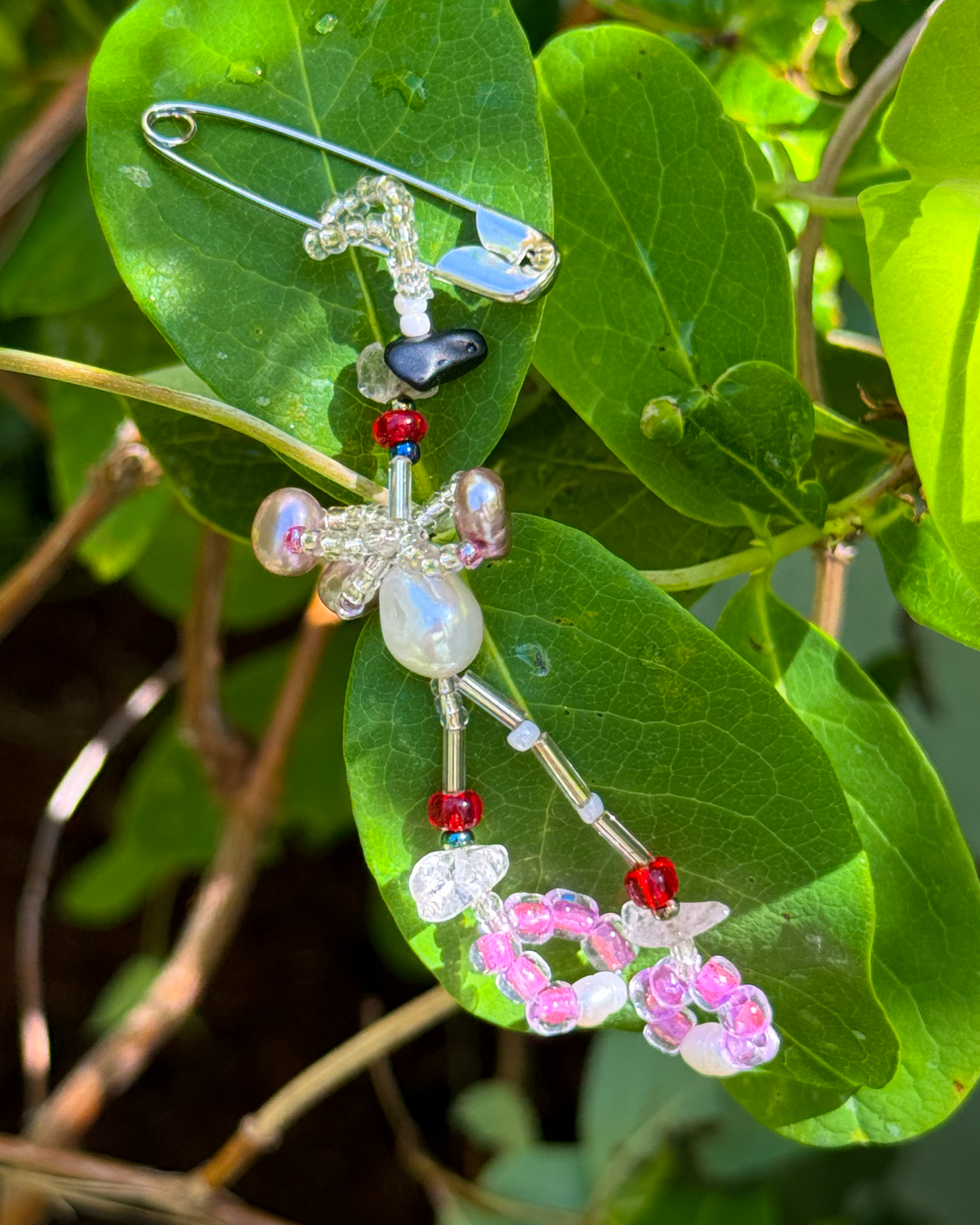A handmade beaded safety pin brooch with an abstract organic design, featuring iridescent pearls, pink and white beads, and red accents, photographed outdoors on a green leaf.