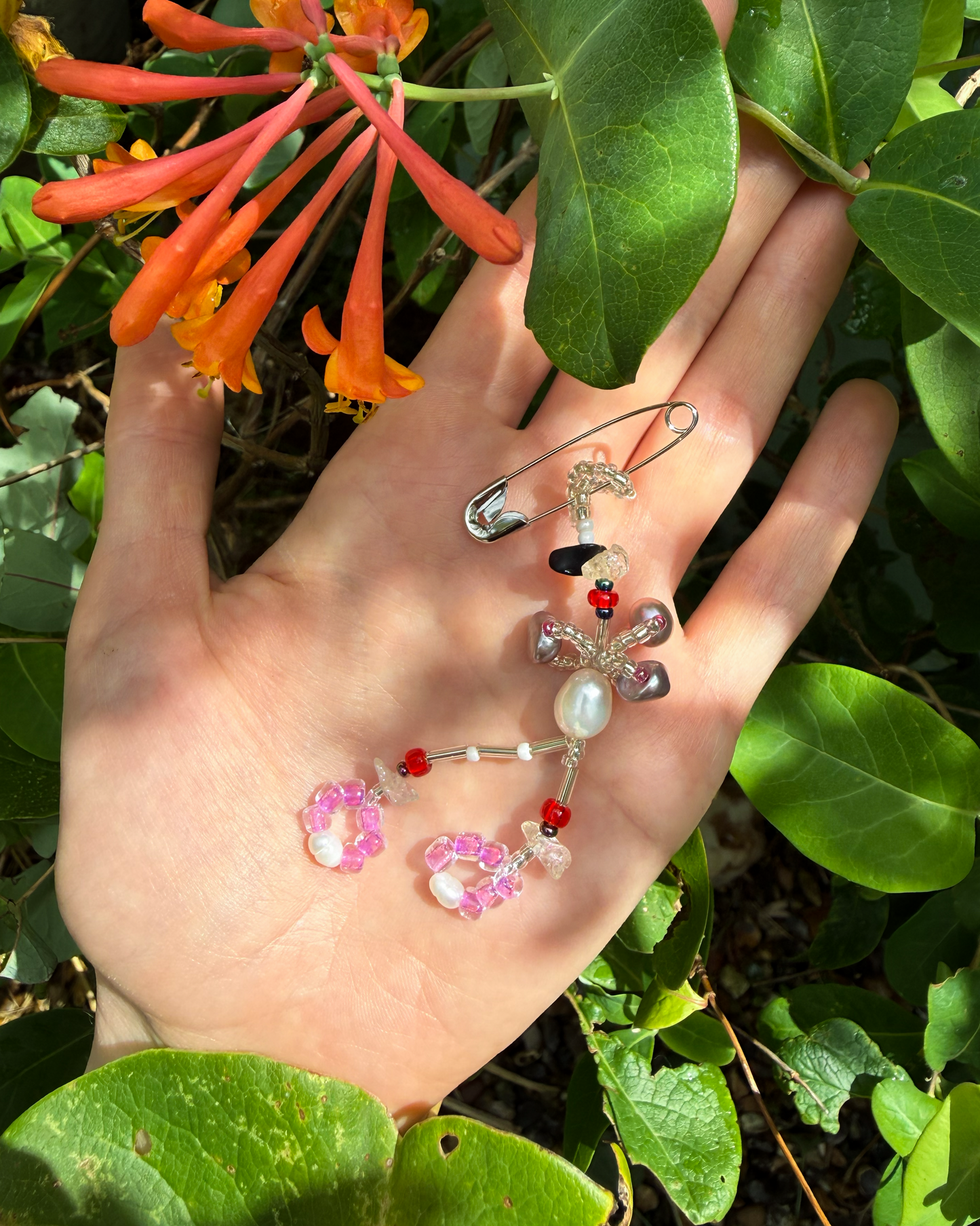A handmade beaded safety pin on an open hand, surrounded by green leaves and orange flowers. The item features a silver safety pin base decorated with a white and lilac pearls, red beads and lilac beads.