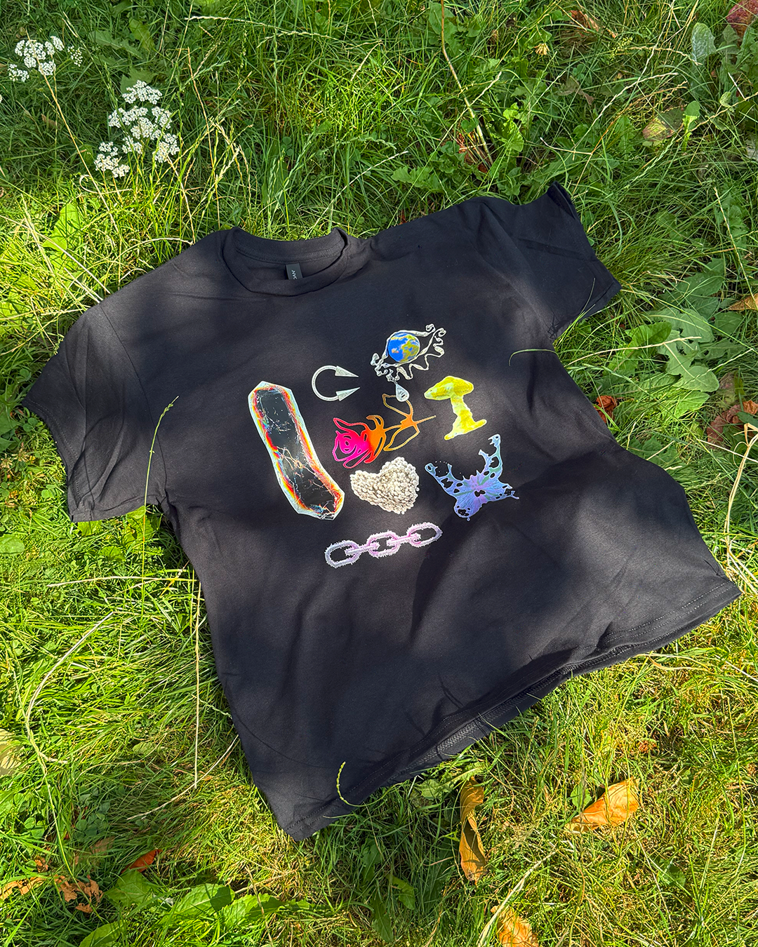 Black t-shirt featuring surreal 3D art: liquid butterfly, chrome chain, and glowing mushroom objects on grass background.