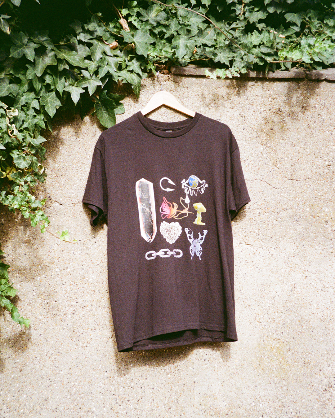 Black t-shirt featuring surreal 3D art: liquid butterfly, chrome chain, and glowing mushroom objects hanging on a wall.