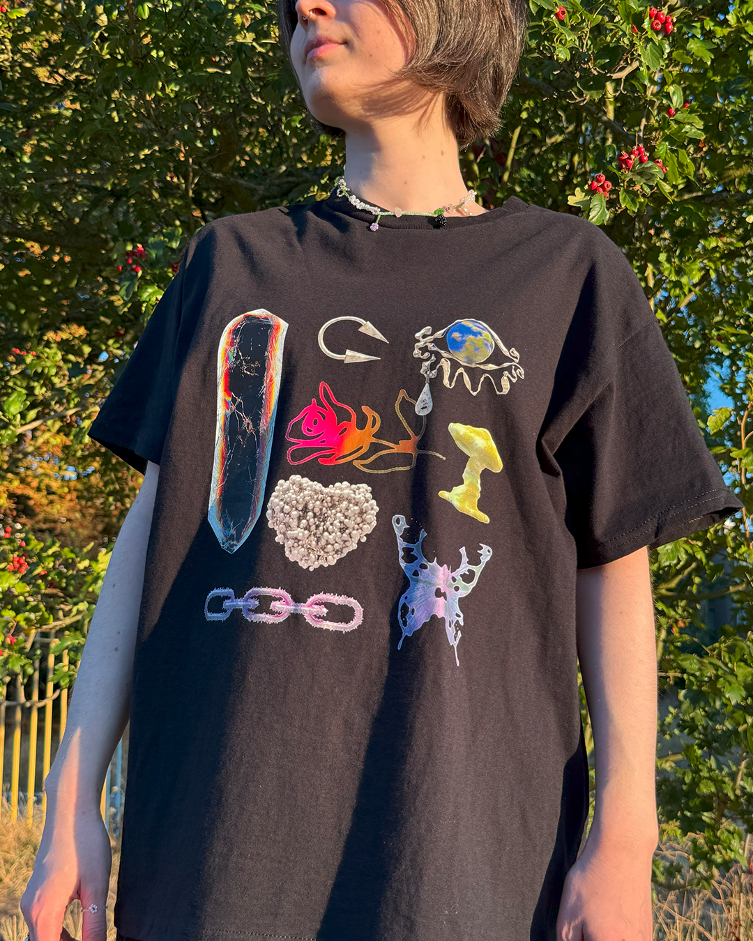 Black t-shirt featuring surreal 3D art: liquid butterfly, chrome chain, and glowing mushroom objects on worn by a woman in the park.